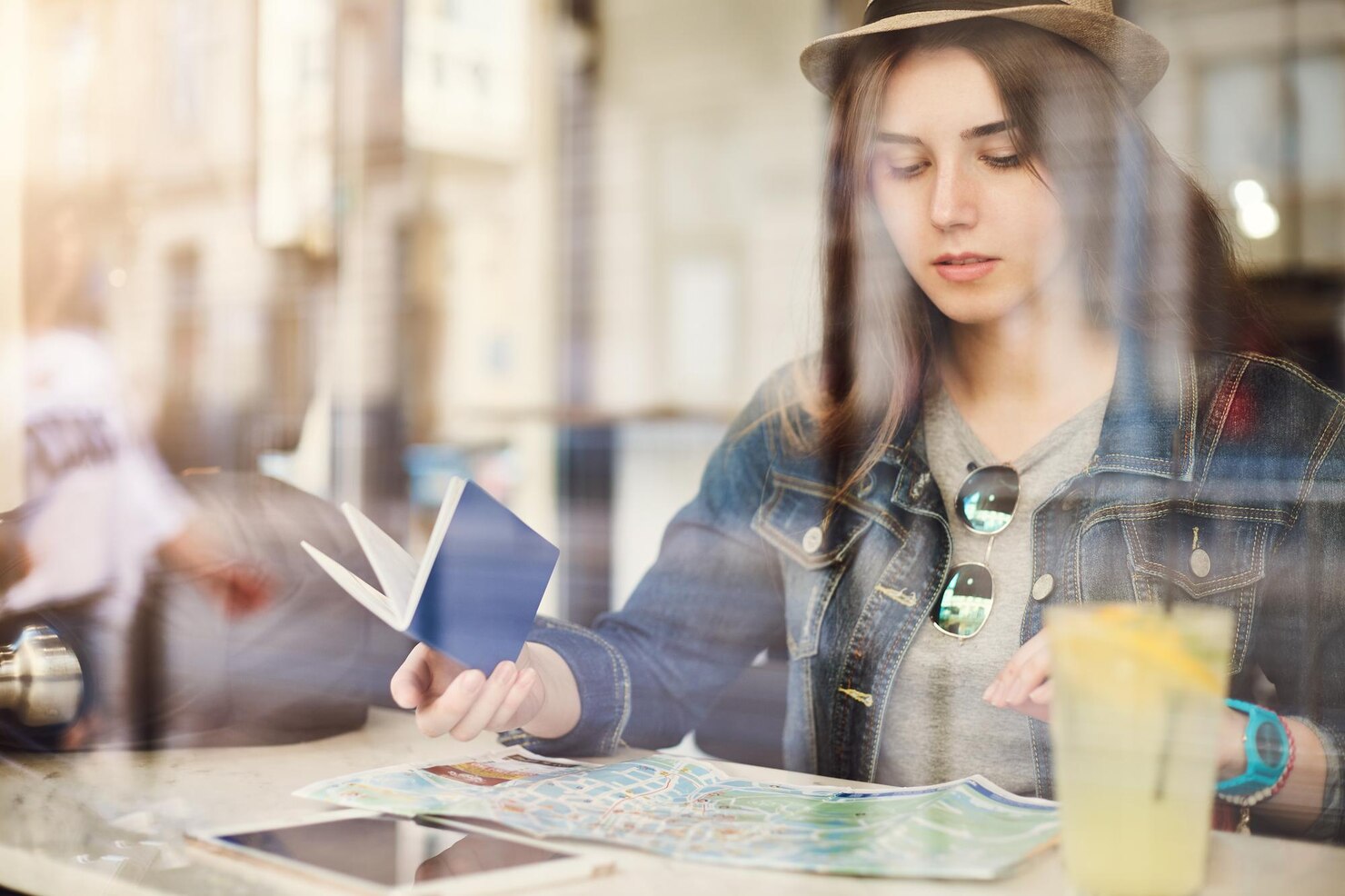 tourist-sitting-cafe-reading-map-holding-passport-drinking-lemonade-busy-city-shot-through-glass_197531-30768